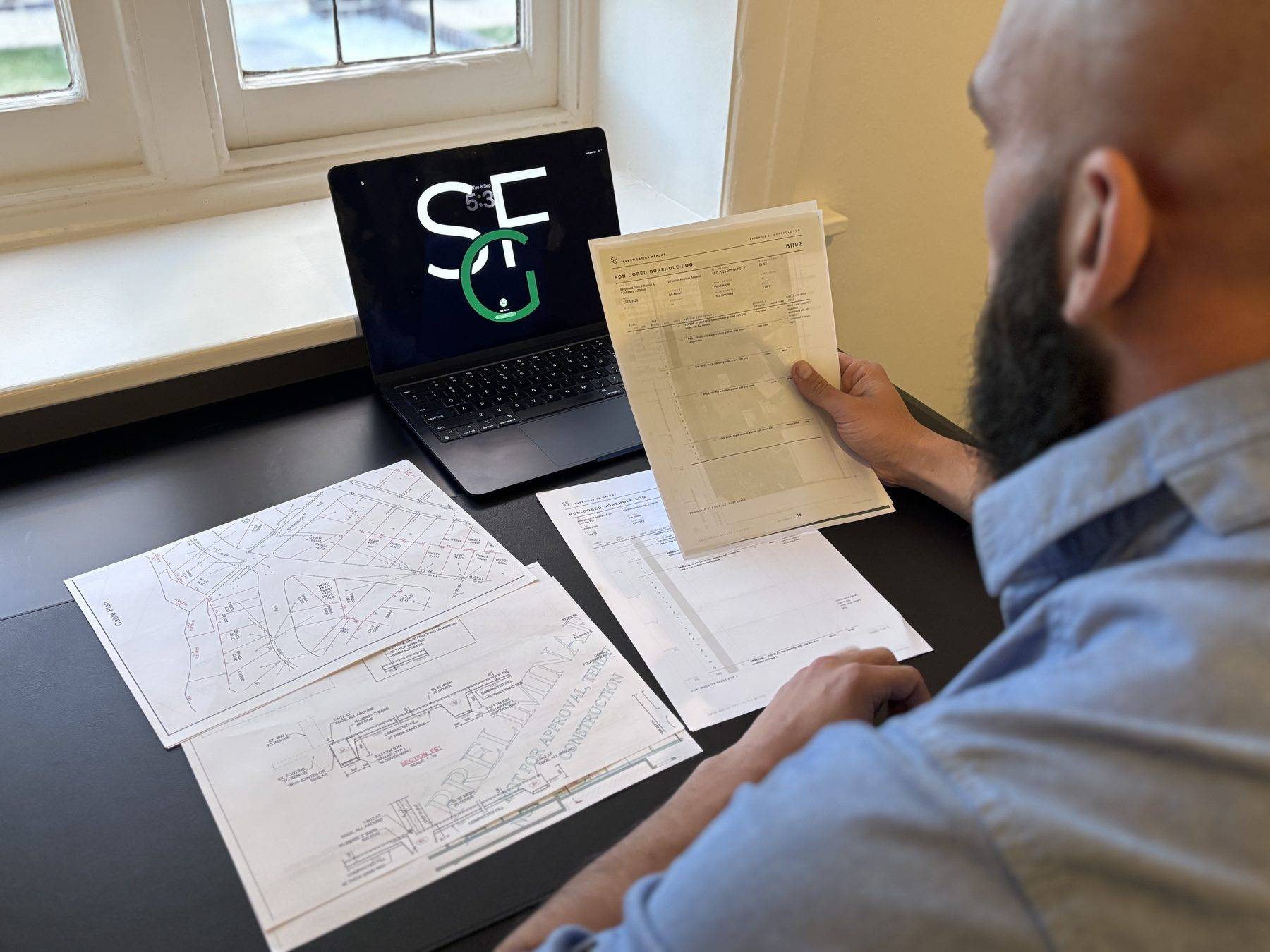 The engineer at the plans desk reading a borehole log against the structural details and a utility plan