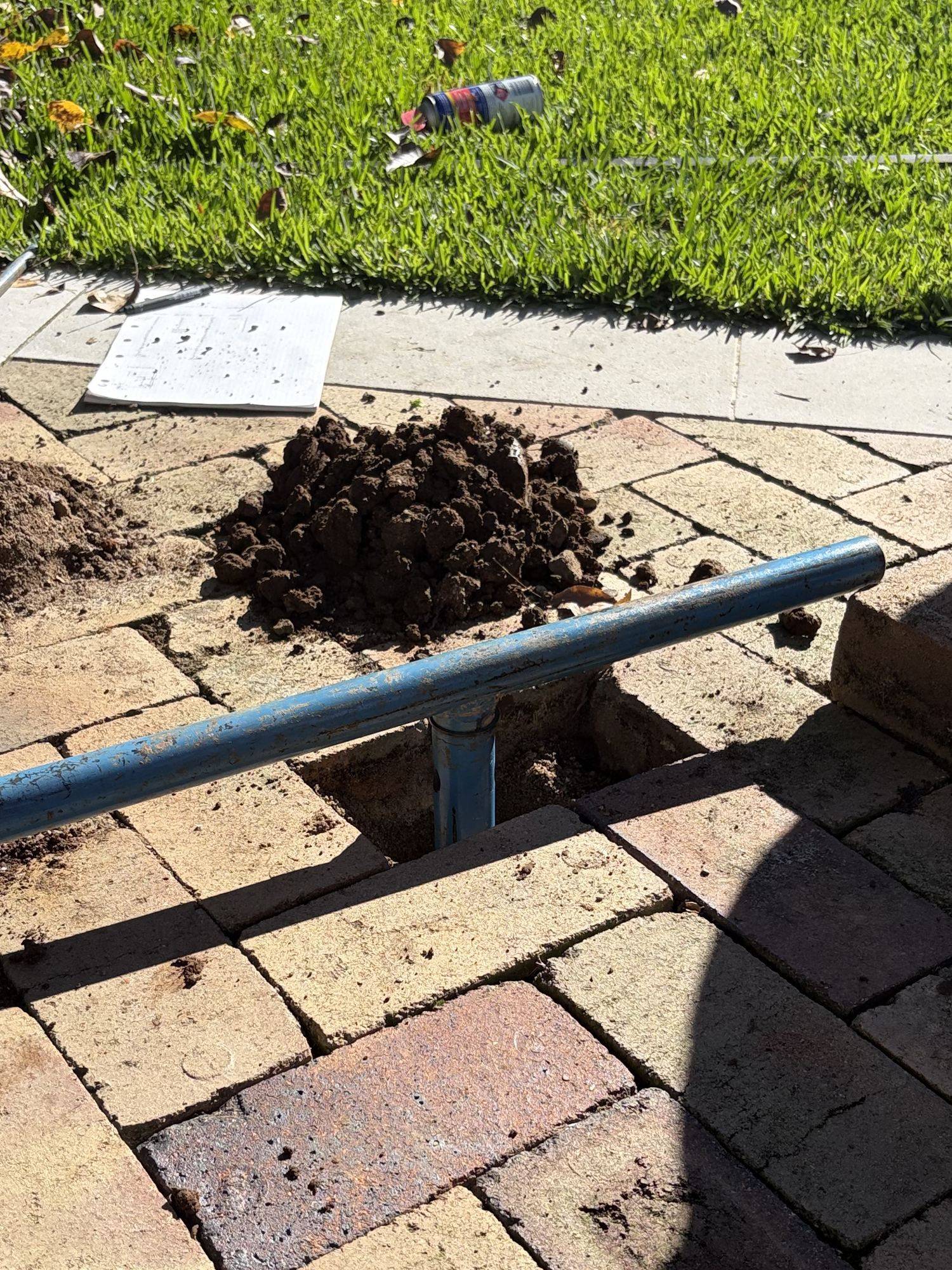 Hand auger boring through lifted pavers in a courtyard where a rig cannot reach, spoil and field log alongside