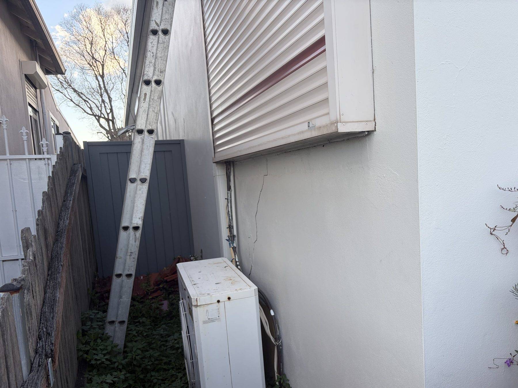 A side passage in Punchbowl barely wider than the air-conditioning unit standing in it, between a timber fence and a rendered wall